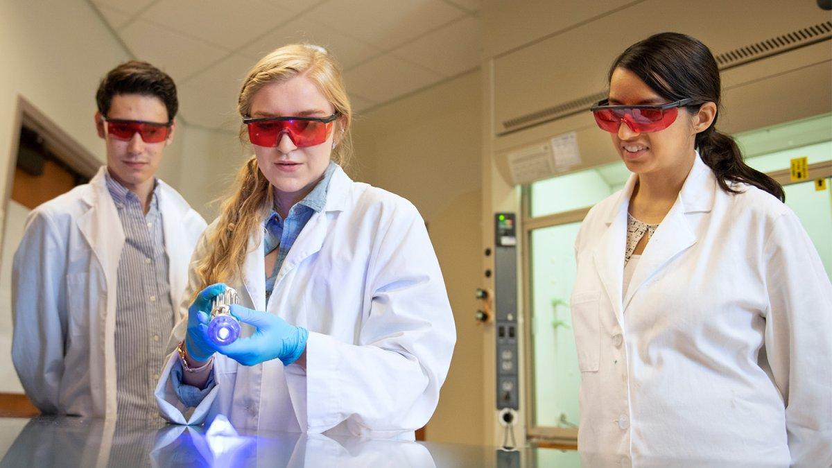 students in lab coats and protective glasses work on an experiment